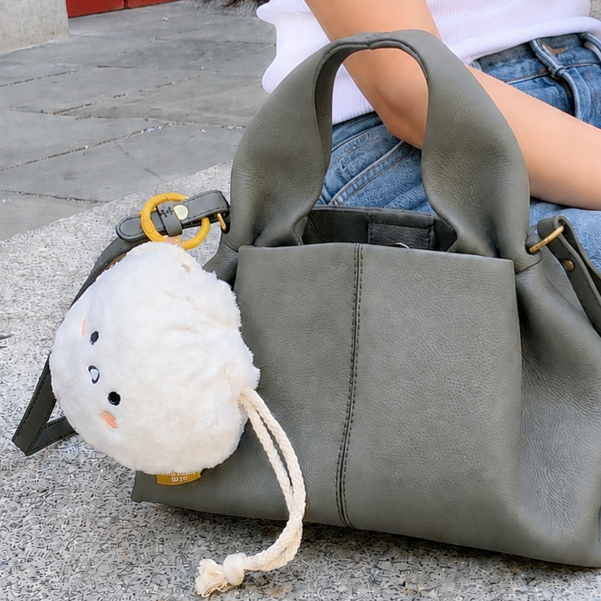 A large olive green faux-leather handbag with a white plush drawstring pouch charm featuring a cartoon face, shown next to a person sitting outdoors on a stone surface.