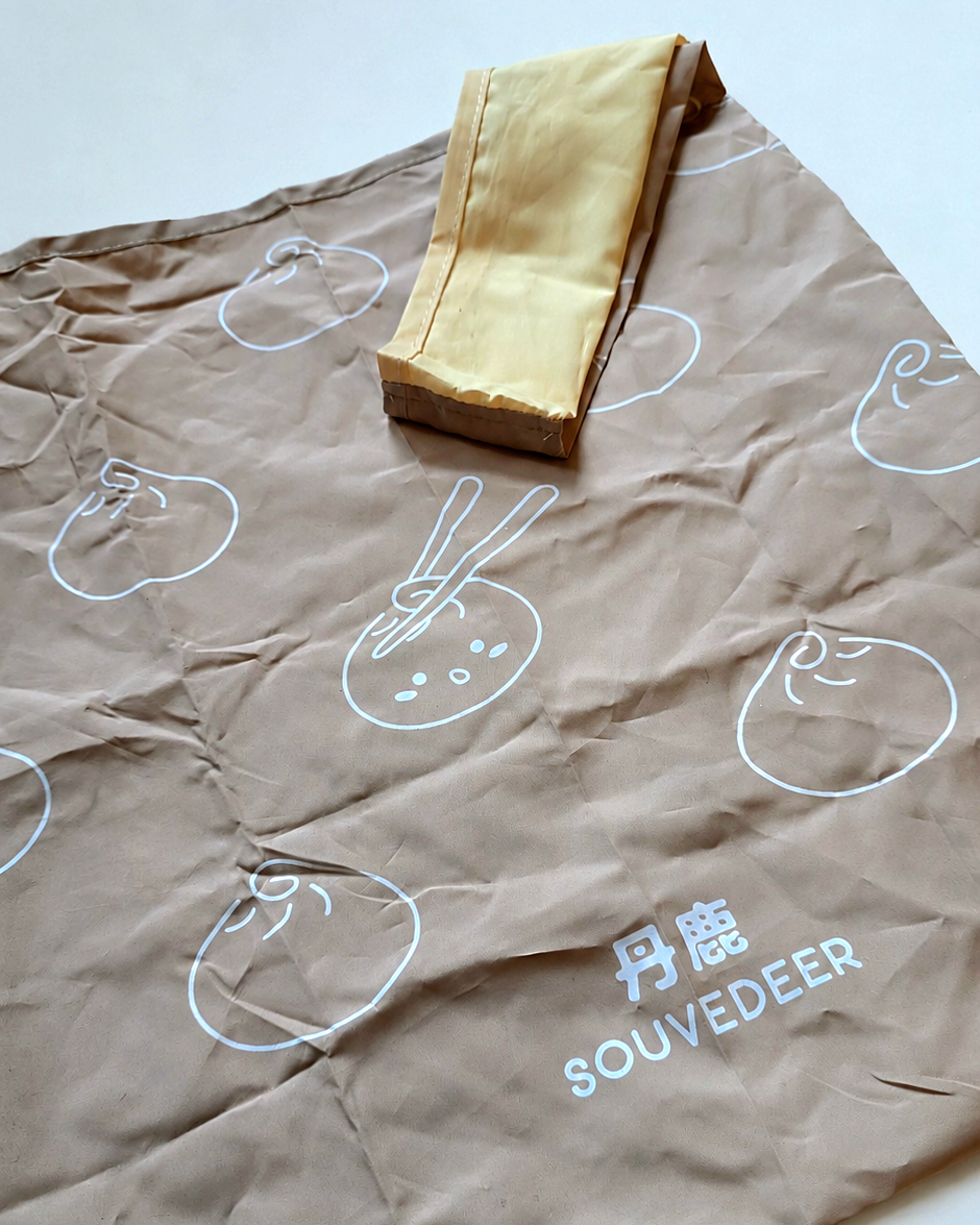 A brown fabric pouch or sleeve with a white dumpling print and the visible brand name 'SOUVEDEER' laid flat on a light background, with a folded gold-colored fabric piece placed on top.